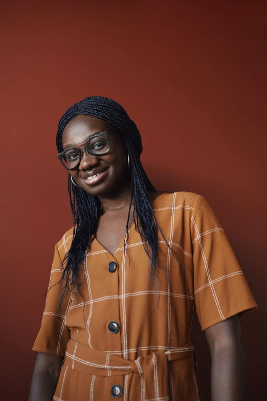 a woman standing in front of a red wall, by Chinwe Chukwuogo-Roy, hurufiyya, wavy long black hair and glasses, wearing a brown, aged 2 5, full frame image