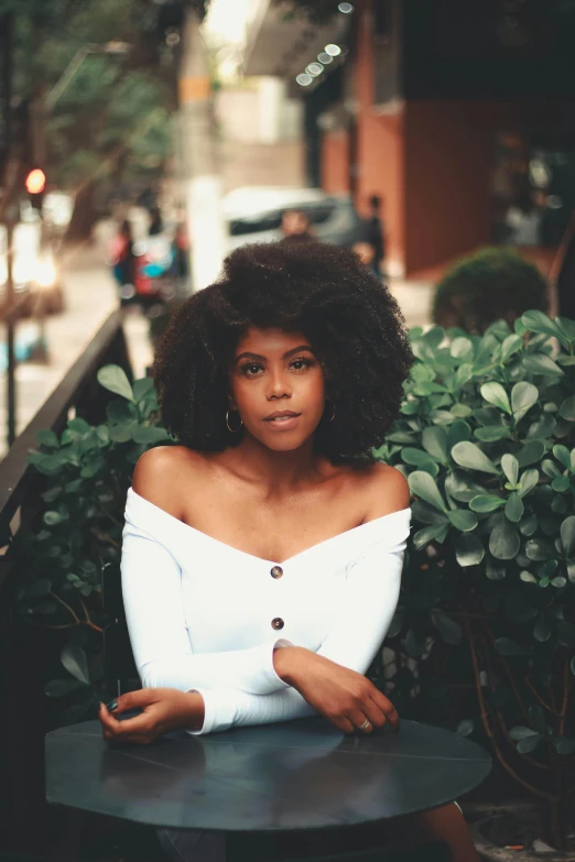 a woman in a white top sitting at a table, by Lily Delissa Joseph, pexels contest winner, big hair, sitting in a tree, black young woman, square