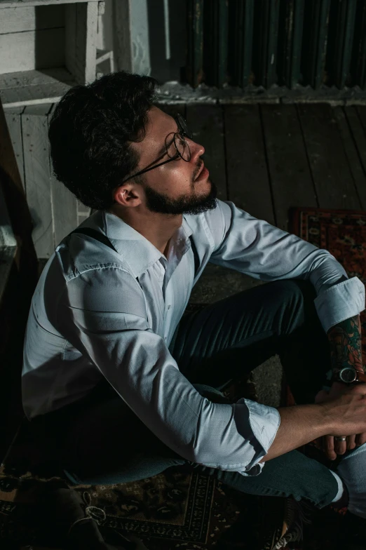 a man sitting on a rug in a room, an album cover, inspired by Camilo Egas, pexels contest winner, jewish young man with glasses, elegant pose, (night), white shirt and jeans