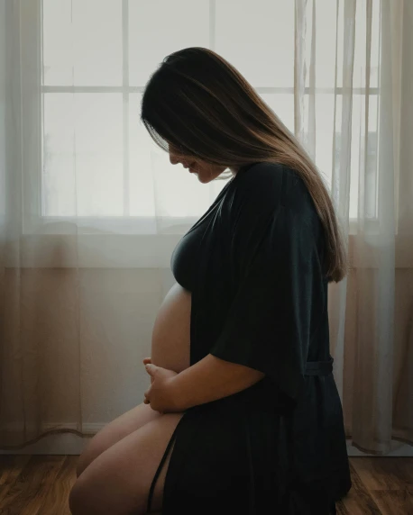 a pregnant woman sitting on the floor in front of a window, pexels contest winner, happening, trans rights, arm around her neck, girl standing, she has a distant expression