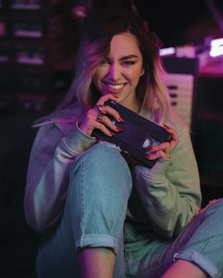 a woman sitting on the ground holding a cell phone, an album cover, inspired by Elsa Bleda, pexels contest winner, happening, purple neon light, taking control while smiling, pokimane, official store photo