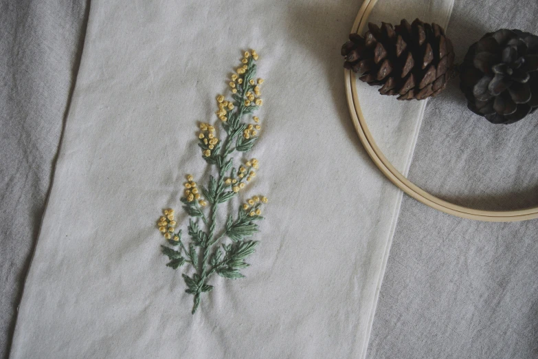 a piece of embroidery sitting on top of a table next to a pine cone, trending on unsplash, yellow flowers, minimalistic aesthetics, wearing gilded ribes, cotton fabric