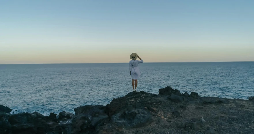 a woman standing on top of a rock next to the ocean, in the evening, wearing a white bathing cap, looking onto the horizon, zac retz