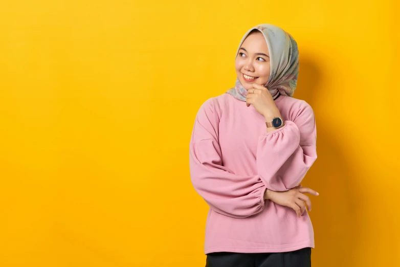 a woman wearing a headscarf standing against a yellow wall, inspired by JoWOnder, shutterstock, hurufiyya, pink shirt, wearing casual sweater, malaysian, smart casual