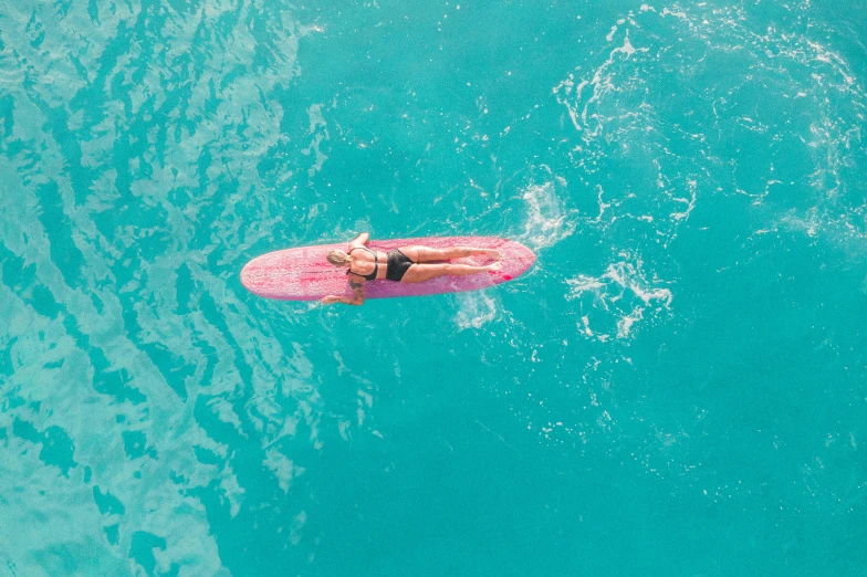 a woman laying on a surfboard in the ocean, pexels contest winner, pink and teal, flat colour, drone photograpghy, pool tubes