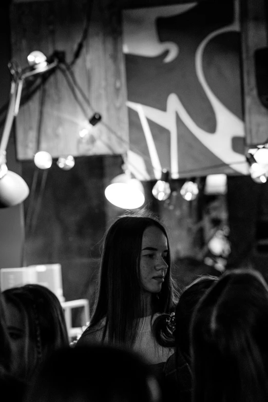 a woman standing in front of a crowd of people, a black and white photo, by Tamas Galambos, unsplash contest winner, process art, mysterious coffee shop girl, evening lighting, girl with long hair, childish look