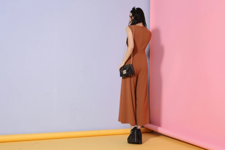 a woman standing in front of a pink and blue wall, inspired by Esaias Boursse, wearing an orange jumpsuit, dark brown duster, rear-shot, brown:-2