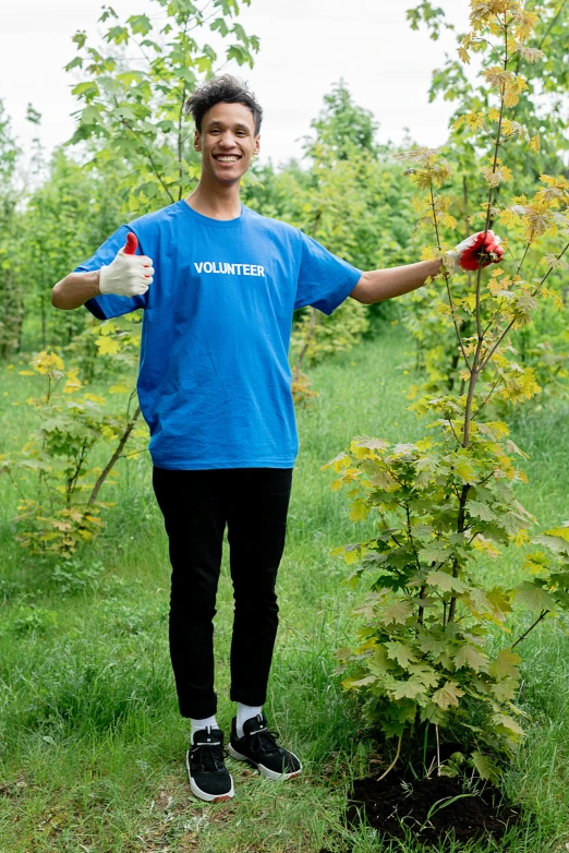 a man in a blue shirt standing next to a tree, giving a thumbs up, connected to nature via vines, wearing a t-shirt, jugendstill