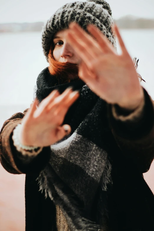 a woman standing in front of a body of water, an album cover, by Tobias Stimmer, trending on pexels, visual art, fingerless gloves, ginger hair and fur, waving hands, face is wrapped in a black scarf