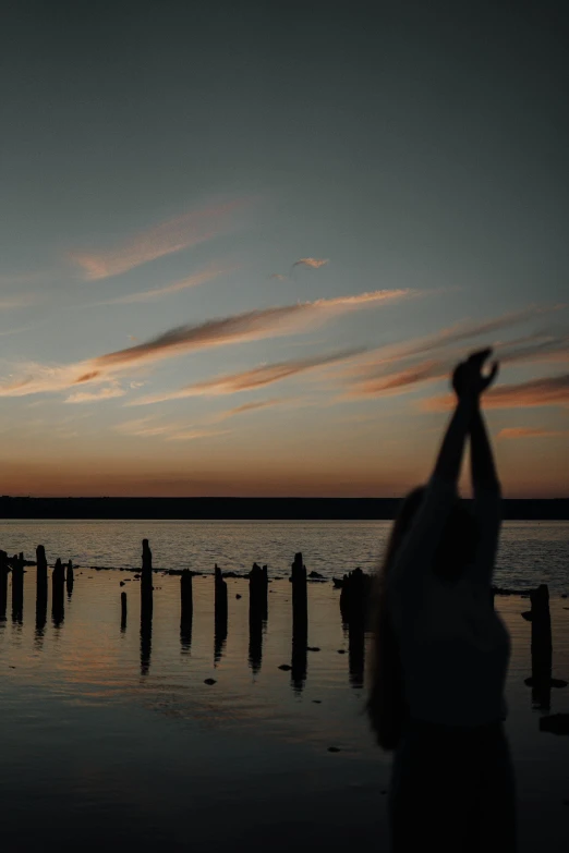 a man standing on top of a beach next to a body of water, a picture, unsplash, happening, hands reaching for her, spring evening, low quality footage, shrugging arms