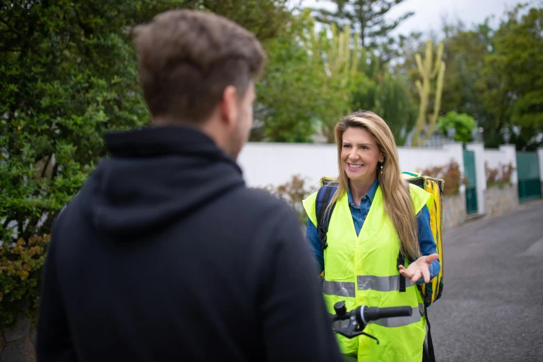 a woman in a yellow vest talking to a man in a black hoodie, delivering mail, lachlan bailey, high quality product image”, high resolution image