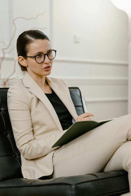 a woman sitting in a chair reading a book, wearing a suit and glasses, mental health, giving an interview, intense knowledge