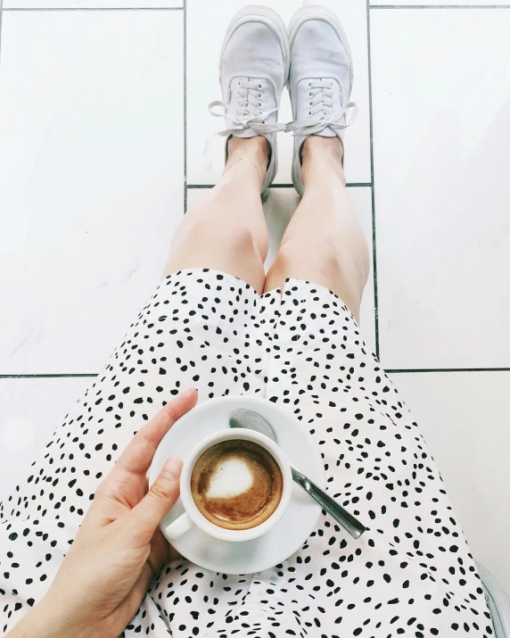 a person sitting on a tiled floor holding a cup of coffee, by Lucia Peka, instagram picture, white with black spots, sneaker photo, in a dress