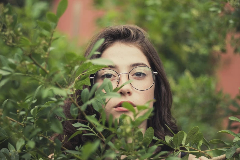 a woman wearing glasses standing in front of a bush, inspired by Elsa Bleda, pexels contest winner, hyperrealism, square glasses, teenager girl, green foliage, charli bowater