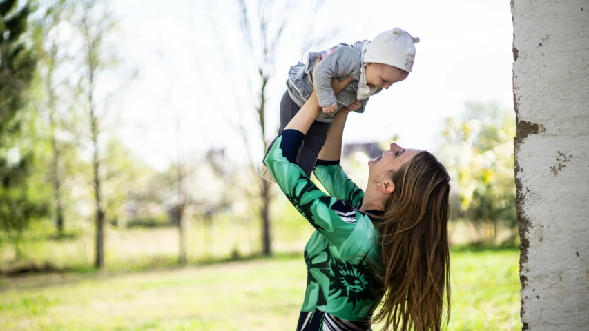 a woman holding a baby up in the air, unsplash, happening, green clothes, walking at the park, square, high resolution