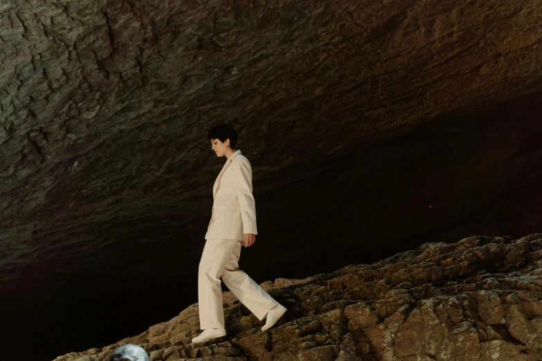 a man in a white suit walking on a rock, an album cover, unsplash, renaissance, declan mckenna, natural cave wall, finn wolfhard, subject detail: wearing a suit