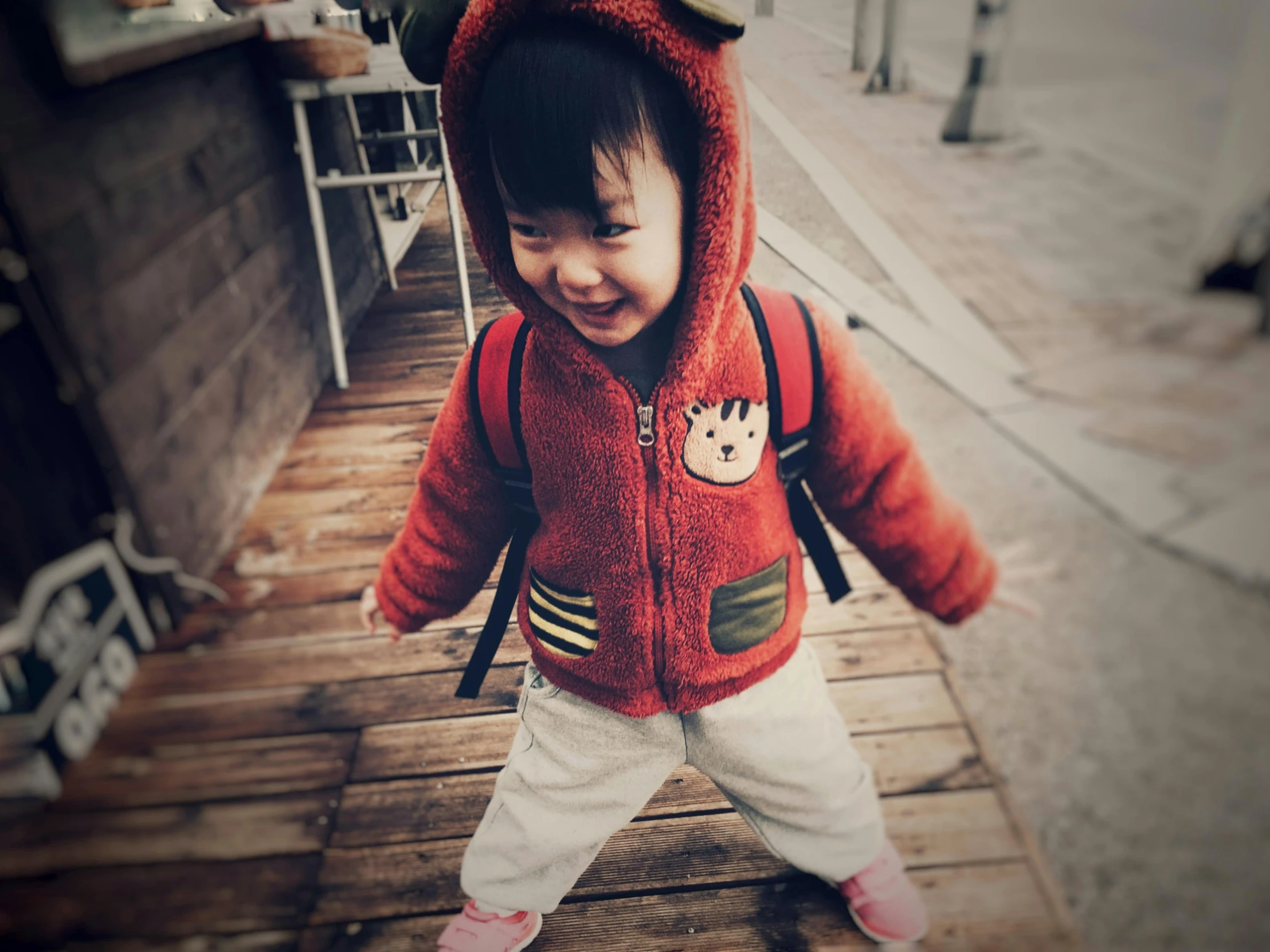 a little girl that is standing on a wooden platform, wearing a red hoodie, li zixin, with a backpack, on a street