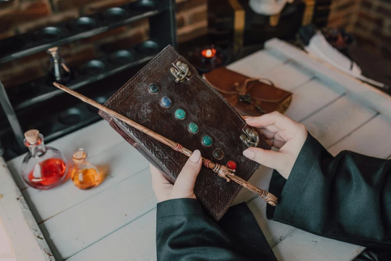 a person holding a book on top of a table, inspired by Fuller Potter, holding the elder wand, gemstones and treasures, leather robes, thumbnail