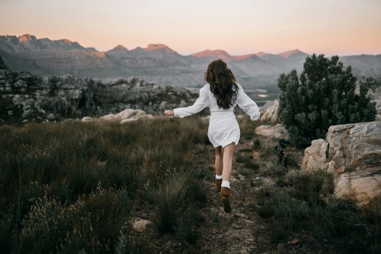 a woman running in a field with mountains in the background, pexels contest winner, wearing a cute white dress, dusk setting, standing on rocky ground, manuka