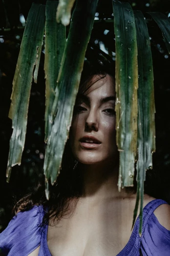 a woman in a blue dress standing under a palm tree, a colorized photo, inspired by Elsa Bleda, pexels contest winner, renaissance, portrait of megan fox, covered in plants, light falling on face, charli xcx