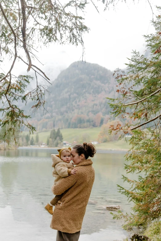 a woman holding a baby near a body of water, girl of the alps, cozy setting, conde nast traveler photo, tan