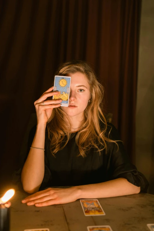 a woman sitting at a table holding a card in front of her face, a portrait, by Julia Pishtar, pexels contest winner, renaissance, in front of a big moon, sydney sweeney, holding an ace card, deity)