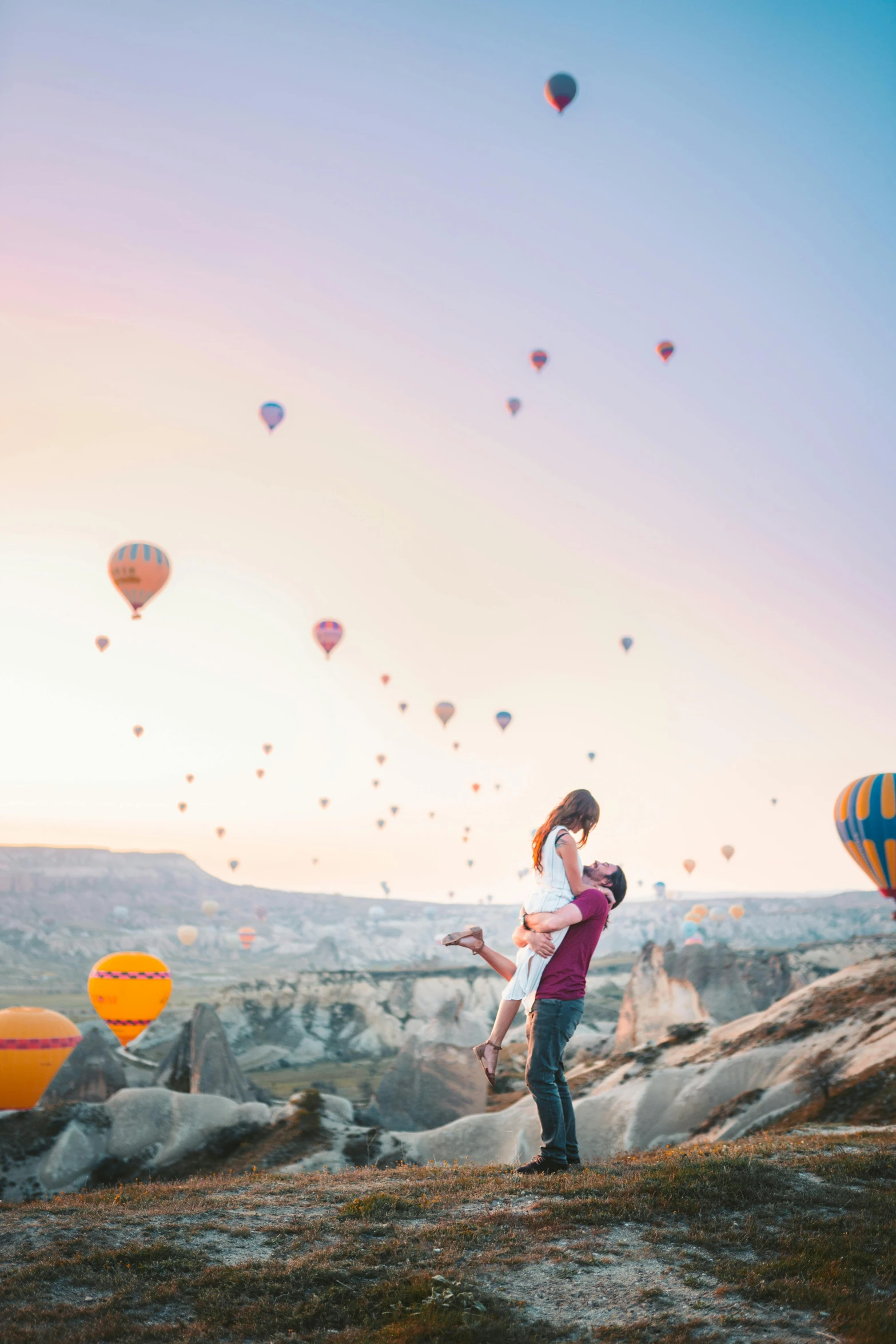 a couple standing on top of a hill surrounded by hot air balloons, cliffs, color photography, please, cinematic”