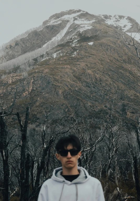 a man wearing sunglasses standing in front of a mountain, by Pablo Rey, discord profile picture, wonbin lee, with dark trees in foreground, chilly dark mood