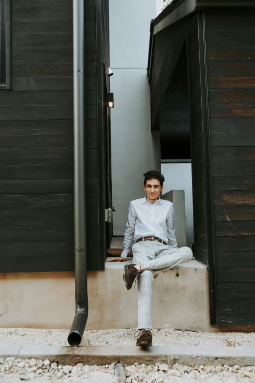 a man sitting on a ledge in front of a building, androgynous person, david marquez, wearing a linen shirt, dark and white