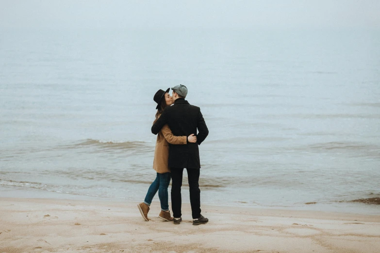 a couple standing next to each other on a beach, pexels contest winner, romanticism, plain background, couple kissing, winter, brown
