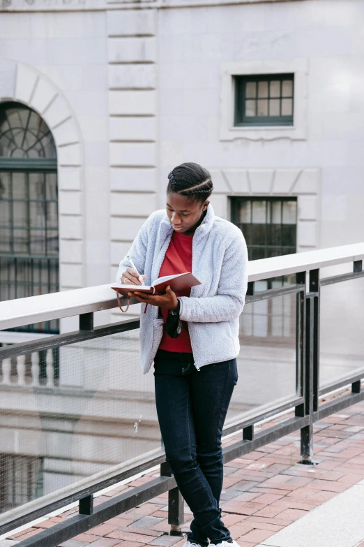 a woman standing on a balcony reading a book, trending on unsplash, academic art, pen and paper, red sweater and gray pants, african american young woman, holding notebook