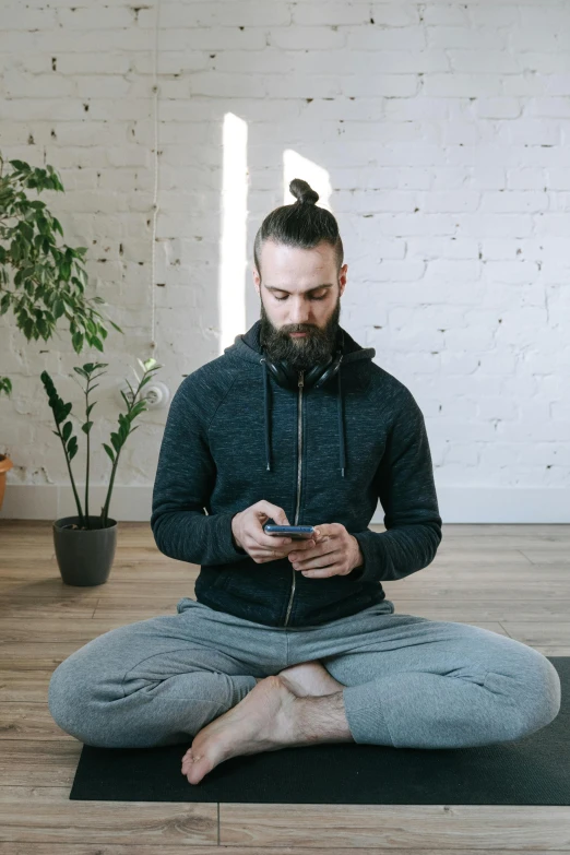 a man sitting on a yoga mat looking at his phone, by Adam Marczyński, trending on pexels, circle beard, wearing gaming headset, centered full body, hair styled in a bun