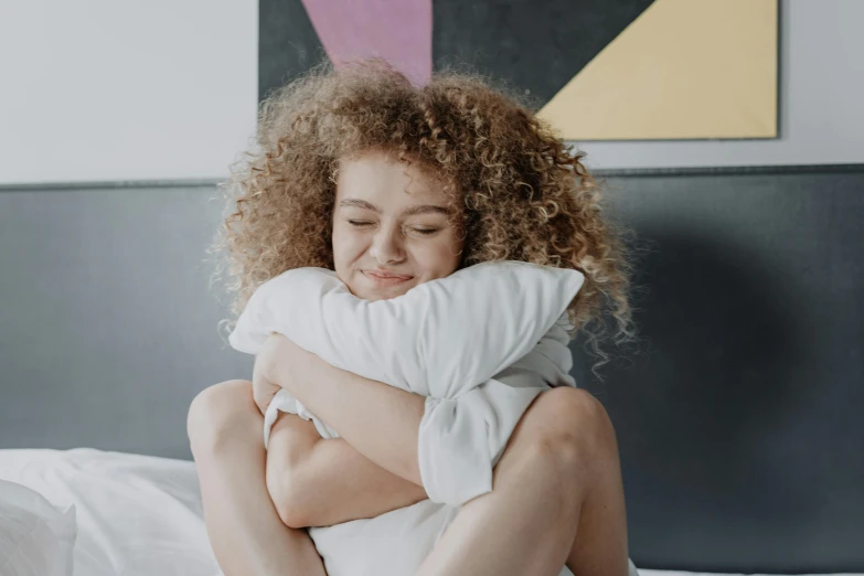 a woman sitting on a bed hugging a pillow, trending on pexels, happening, curly haired, rated t for teen, small smile, slightly pixelated