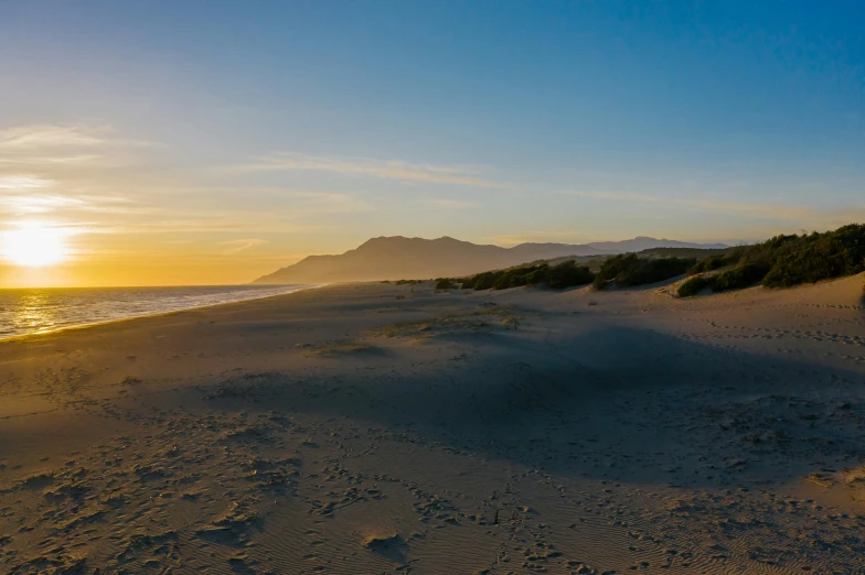 a man riding a surfboard on top of a sandy beach, sun rises between two mountains, hollister ranch, which shows a beach at sunset, dunes