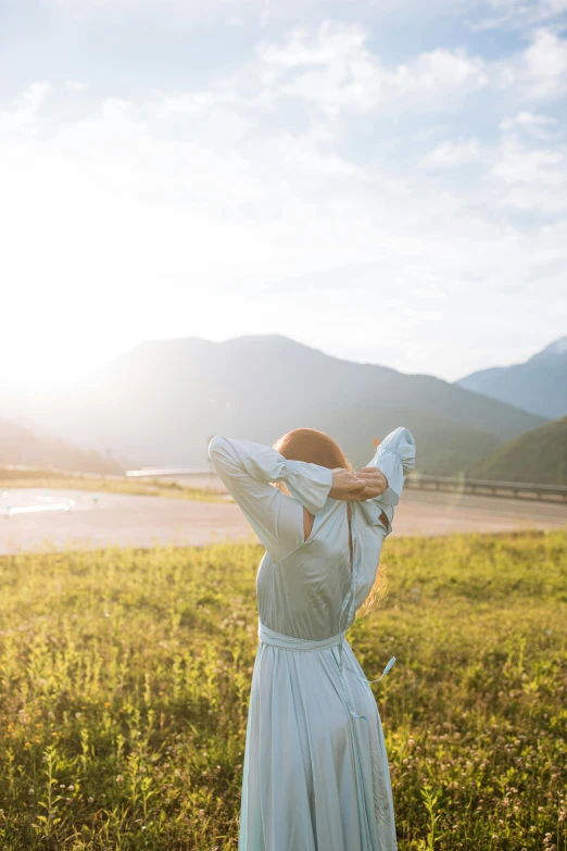 a woman in a white dress standing in a field, trending on unsplash, happening, with mountains in background, sun lit, facing away, playful pose