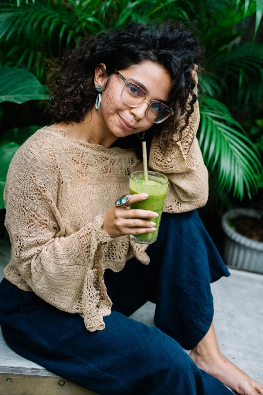 a woman sitting on a bench with a drink in her hand, wearing a green sweater, plants in glasses, olive skin color, moringa juice