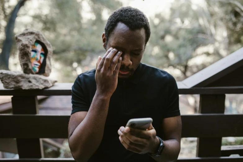 a man sitting on a bench looking at his cell phone, trending on pexels, wet eye in forehead, man is with black skin, devastated, third eye in middle of forehead