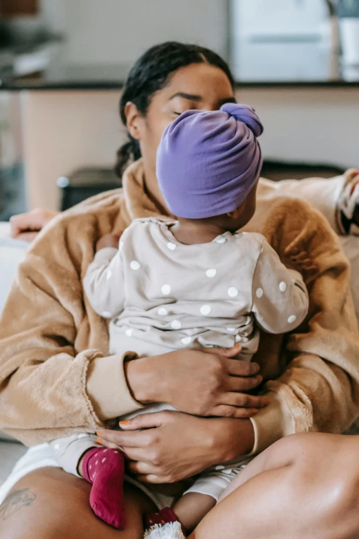 a woman sitting on a couch holding a baby, pexels contest winner, incoherents, wearing a purple cap, manuka, soft and warm, topknot