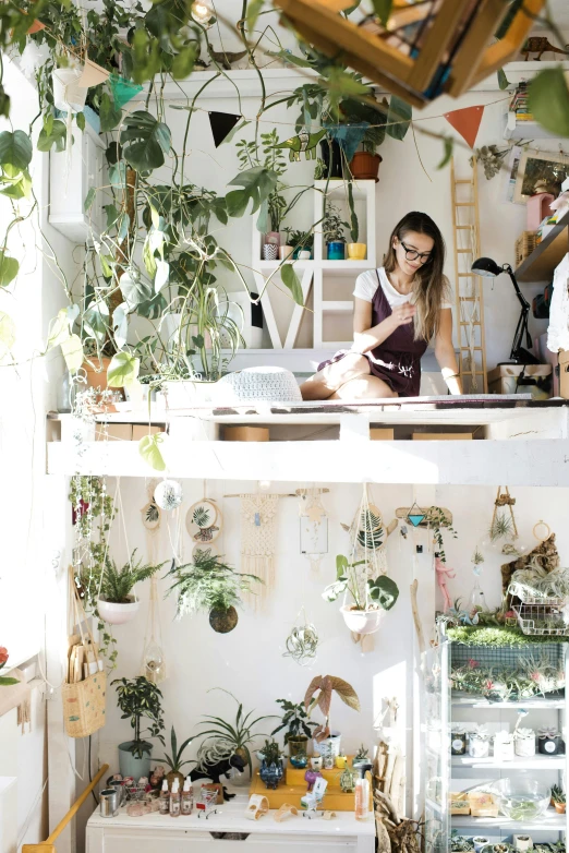 a woman sitting at a desk in a room filled with plants, a photo, trending on pexels, process art, in a white boho style studio, high angle, green house, knick - knacks