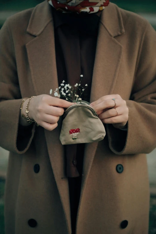 a woman in a brown coat is holding a purse, a cross stitch, by Lucia Peka, pexels contest winner, small crown, handwritten, streetwear, gold coins