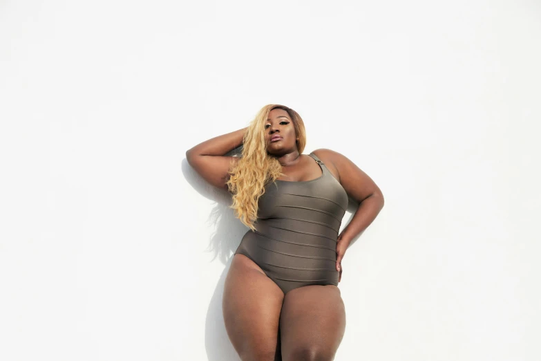 a woman in a bodysuit leaning against a wall, an album cover, by Nyuju Stumpy Brown, unsplash, obese, swimsuit, light-brown skin, blonde goddess