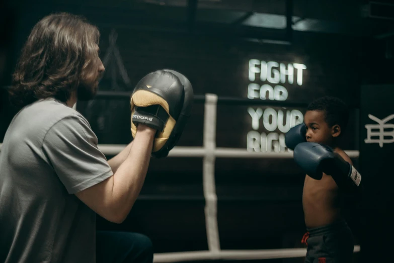 a man standing next to a boy in a boxing ring, by Emma Andijewska, pexels contest winner, symbolism, holding it out to the camera, advert, toddler, glowing