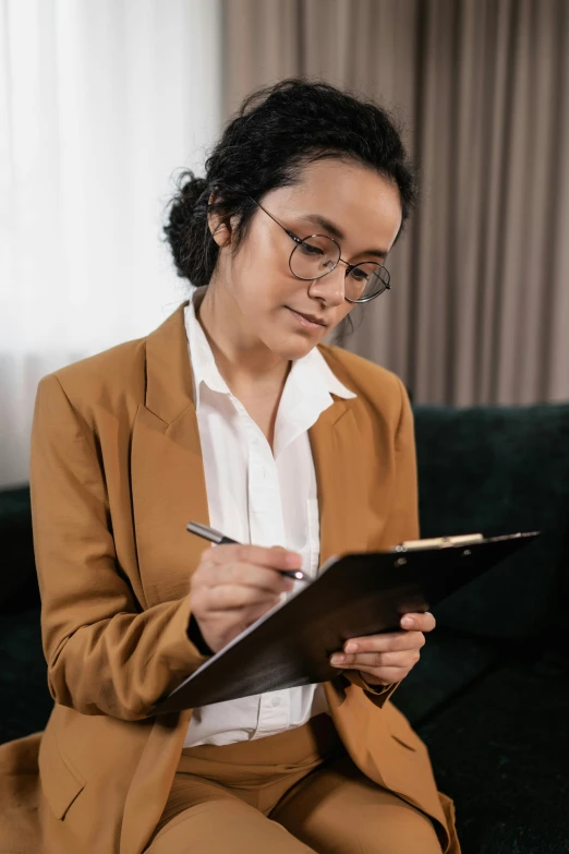 a woman sitting on a couch writing on a clipboard, by Robbie Trevino, tan suit, thumbnail, academic clothing, woman