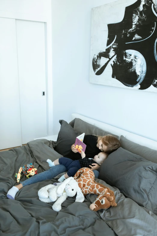a little girl laying on top of a bed next to a stuffed giraffe, visual art, dwell, grey, apartment, reading nook
