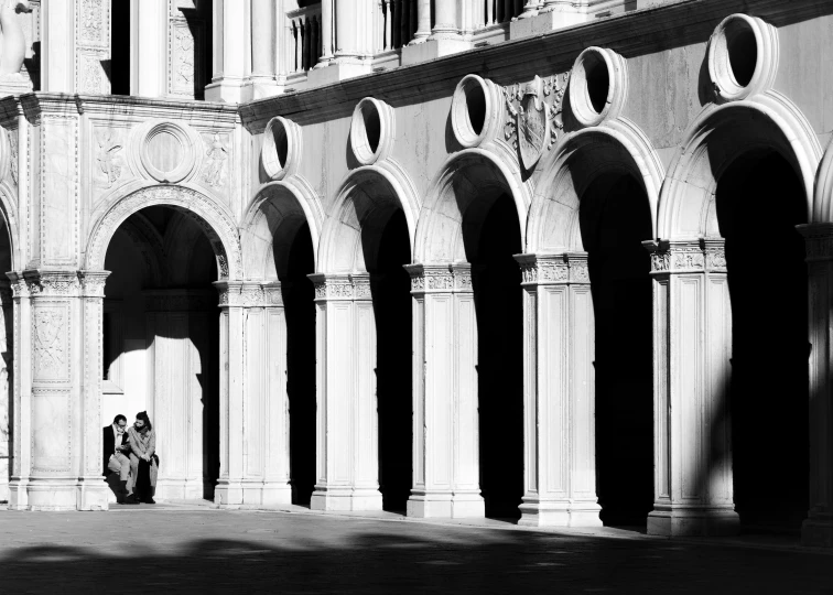 a couple of people that are standing in front of a building, a black and white photo, by Patrick Pietropoli, pexels contest winner, neoclassicism, dry archways, afternoon sunlight, ravnica, arcs