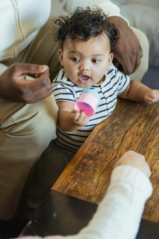 a baby sitting on top of a wooden table, cups and balls, customers, holding a ball, drinking cough syrup