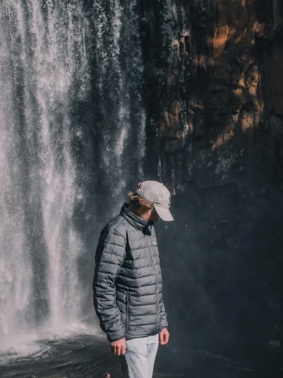 a man standing in front of a waterfall, a picture, by Jan Tengnagel, hat and hoodie, low quality photo, model wears a puffer jacket, view(full body + zoomed out)