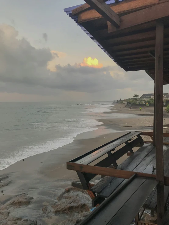 a bench sitting on top of a beach next to the ocean, while it's raining, from the roof, sunset beach, looking towards the camera