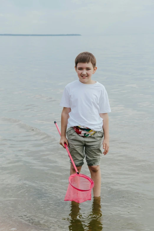 a young boy standing in the water with a pink net, wearing shorts and t shirt, wearing a fisher 🧥, mini model, zoomed in