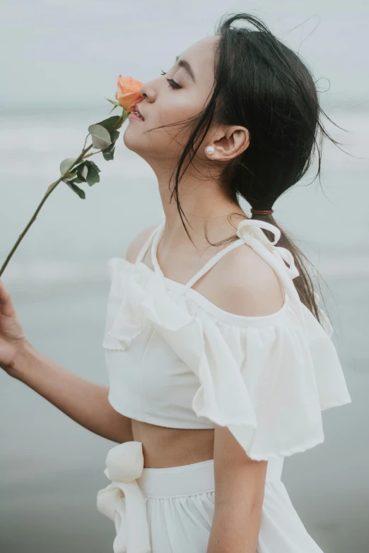 a woman standing on a beach holding a flower, inspired by Ruth Jên, unsplash, renaissance, croptop, white, frill, asian female